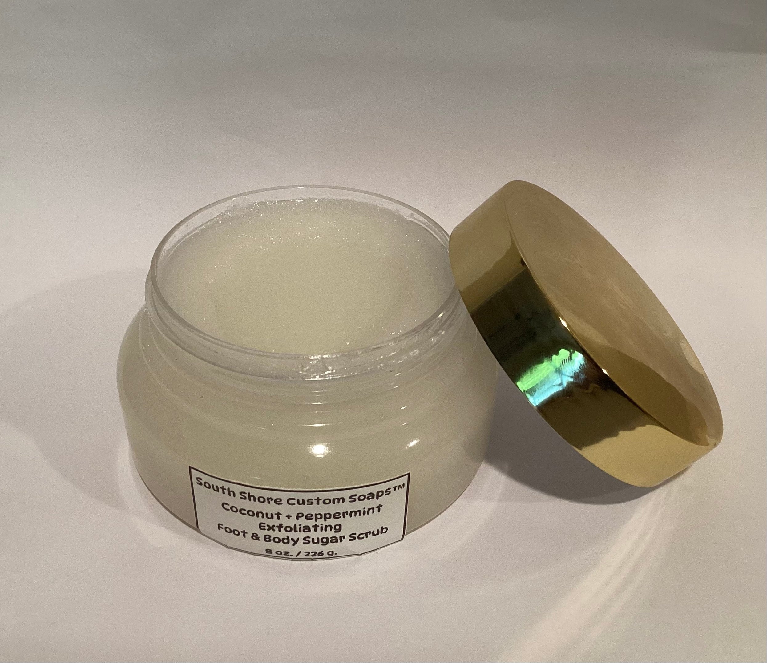 A jar of foot and body sugar scrub with its metallic gold lid leaning against it on a white background. The label reads 'South Shore Custom Soaks Coconut + Peppermint Exfoliating Foot & Body Sugar Scrub'.
