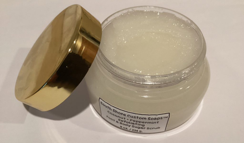 A clear plastic jar of South Shore Custom Soaps coconut and peppermint face & body sugar scrub with a shiny gold lid leaning against it on a white surface.
