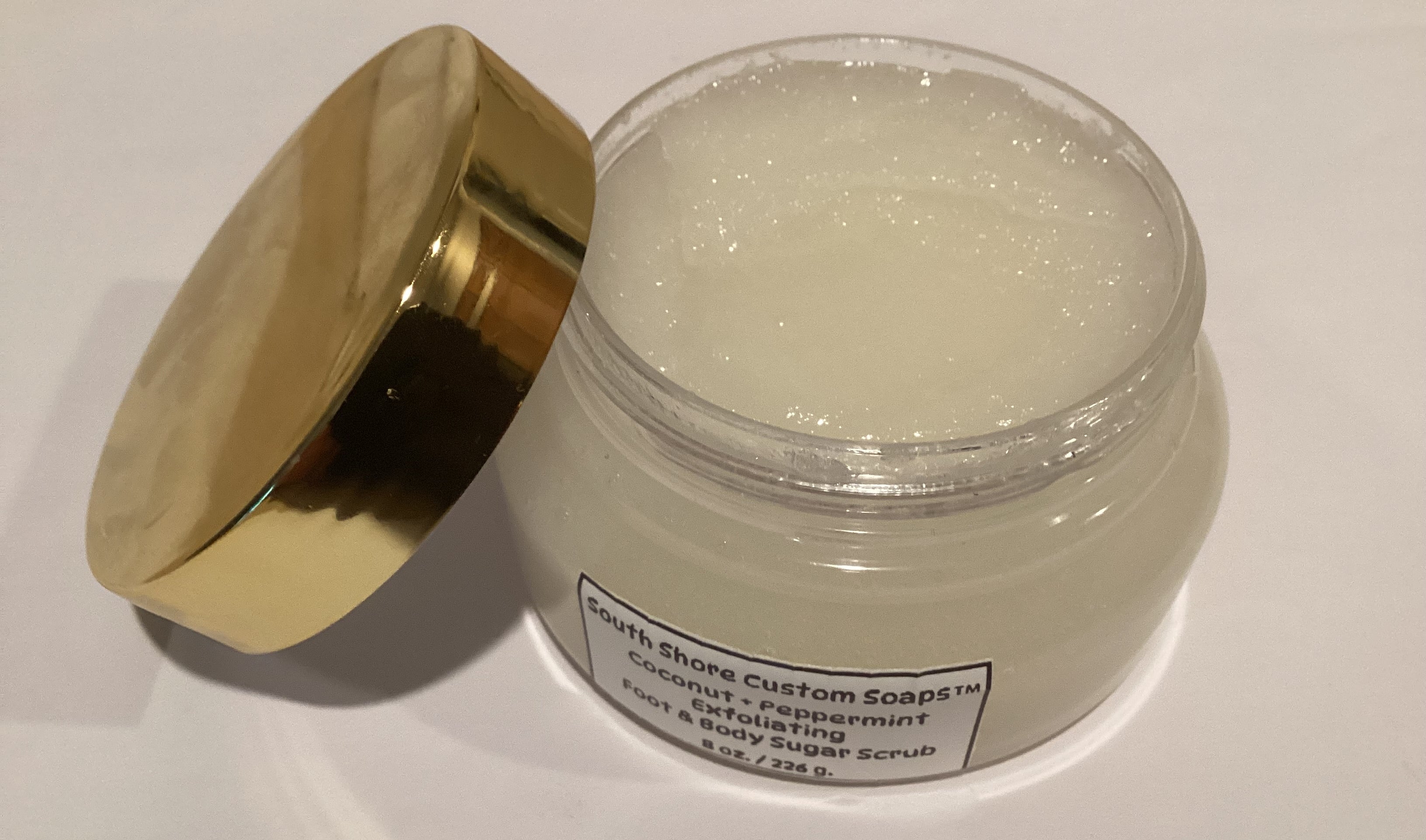 A clear plastic jar of South Shore Custom Soaps coconut and peppermint face & body sugar scrub with a shiny gold lid leaning against it on a white surface.